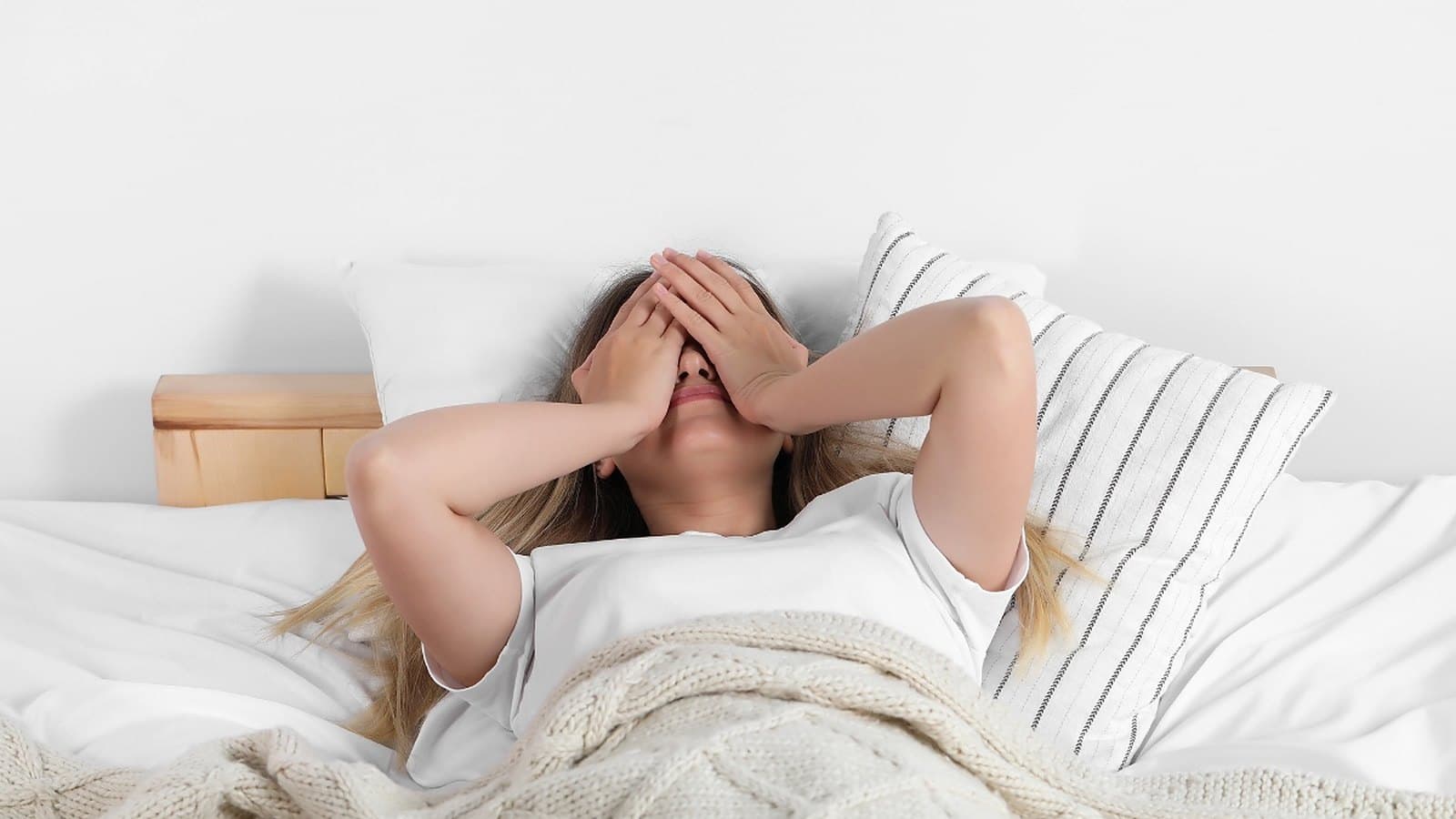 Femme fatiguée au réveil qui se couvre les yeux, allongée dans un lit blanc