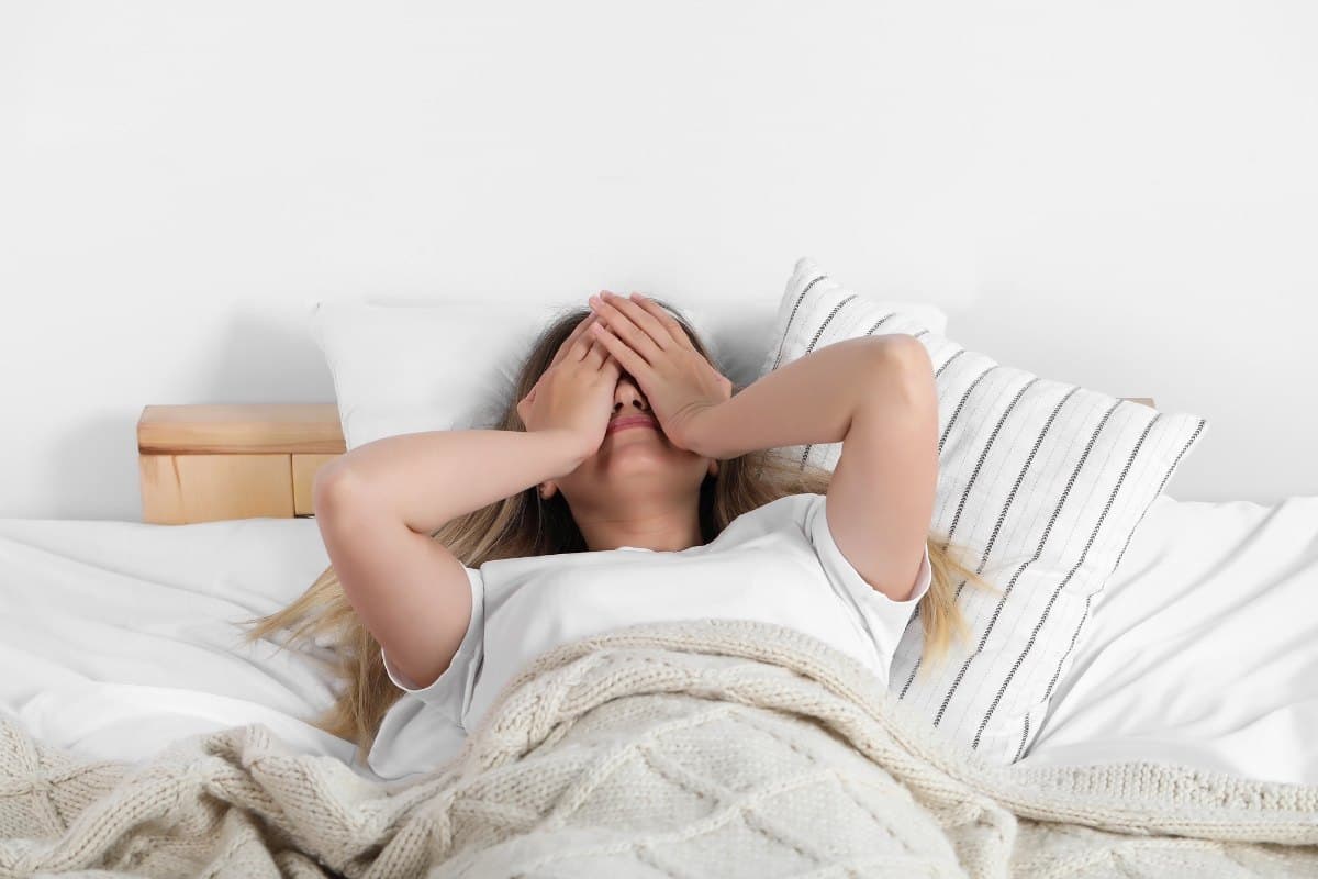 Femme allongée dans son lit, les mains sur le visage, épuisée dès le réveil