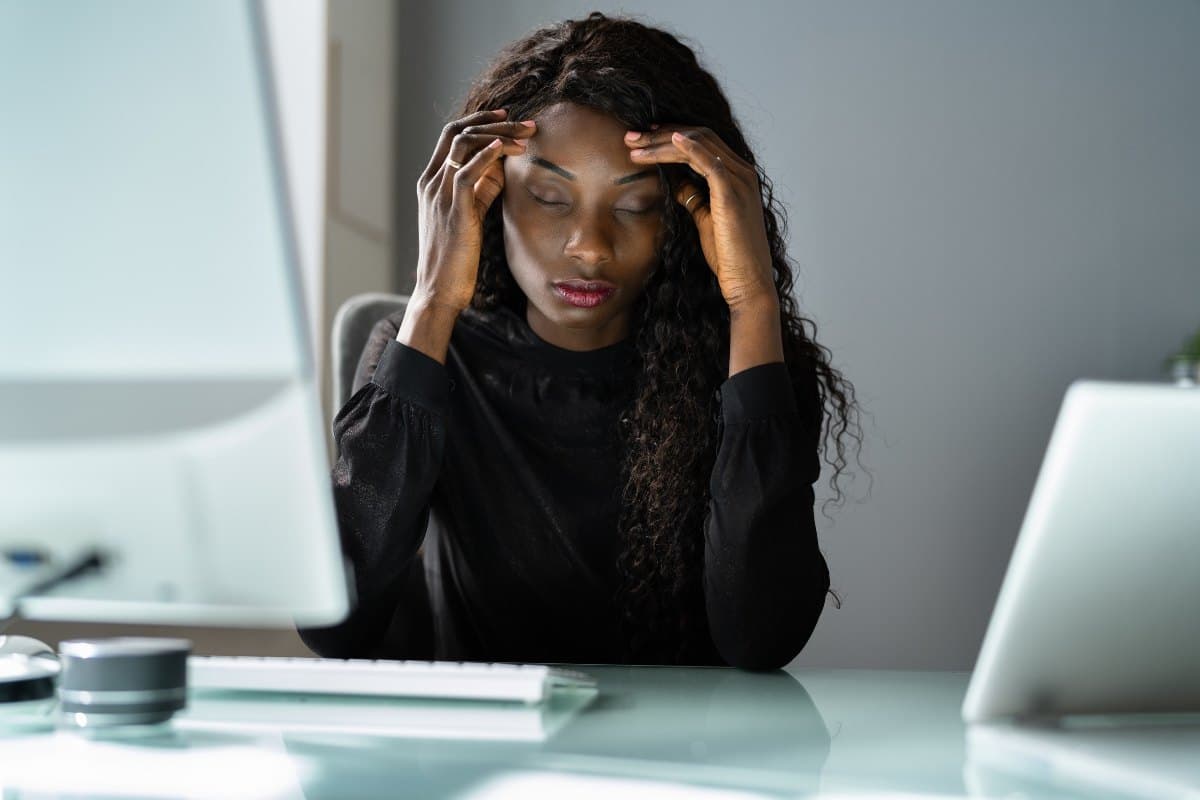 Femme assise à son bureau, les yeux fermés et les mains sur les tempes, submergée par le stress