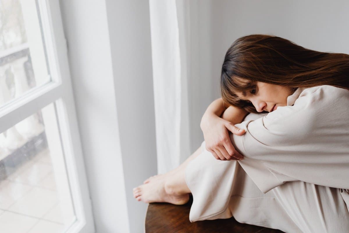 Jeune femme assise près d'une fenêtre, la tête posée sur ses genoux repliés, l'air pensif